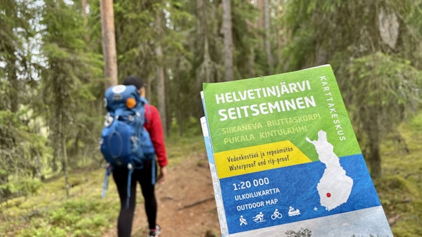 A person wearing a red jacket and a blue backpack is walking along a forest trail. In the foreground, a hand holds a colorful outdoor map labeled 'Helvetinjärvi Seitsemisen' with various details and symbols indicating hiking, cycling, and campfire possibilities. The background is filled with tall green trees and lush vegetation, creating a serene natural setting.