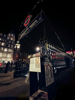 A nighttime urban scene featuring the entrance to Westminster Station, marked by a recognizable sign with the London Underground logo. In the foreground, newspapers are displayed under a light, casting a glow on the metal fence. The surrounding area includes historical buildings and streetlights that enhance the ambiance.