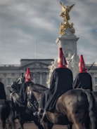 A procession of mounted guards with black horses whose riders wear uniforms with red feathered helmets. They are in front of a grand building that appears to be a palace. In the foreground, the focus is on the riders and their horses, while in the background, a statue with a golden figurine stands prominently atop a plinth, topped with a winged figure.