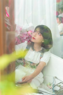 A young person with dark hair sits in a reflective, serene setting surrounded by pink and white flowers. Their gaze is directed upwards, giving an impression of contemplation or wonder. Soft lighting and delicate colors create a gentle and dreamy atmosphere.