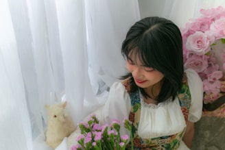 A young woman with dark hair and a floral-patterned dress is holding a bouquet of pink flowers and smiling softly. She is surrounded by white semi-transparent curtains and a large arrangement of pink roses to the side.