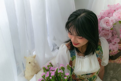A young woman with dark hair and a floral-patterned dress is holding a bouquet of pink flowers and smiling softly. She is surrounded by white semi-transparent curtains and a large arrangement of pink roses to the side.