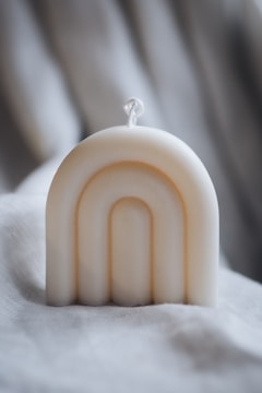 A cream-colored, arch-shaped candle is positioned on a soft, textured fabric background. The candle features a simple rope wick and smooth, minimalist curves that create an elegant design.