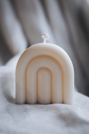 A cream-colored, arch-shaped candle is positioned on a soft, textured fabric background. The candle features a simple rope wick and smooth, minimalist curves that create an elegant design.