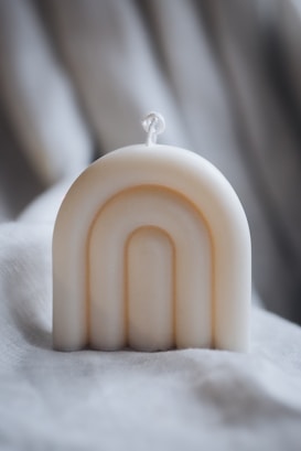 A cream-colored, arch-shaped candle is positioned on a soft, textured fabric background. The candle features a simple rope wick and smooth, minimalist curves that create an elegant design.