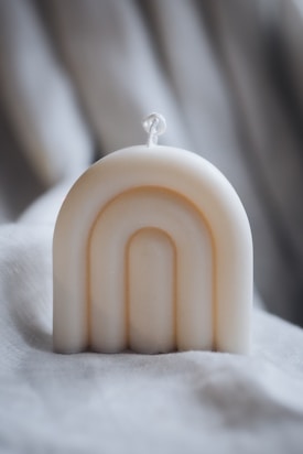 A cream-colored, arch-shaped candle is positioned on a soft, textured fabric background. The candle features a simple rope wick and smooth, minimalist curves that create an elegant design.