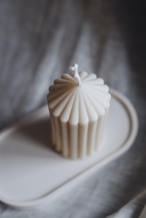 A fluted, cream-colored candle with a wick at the top resting on an oval tray against a blurred gray background. The candle's surface features regular vertical grooves adding a textured look.