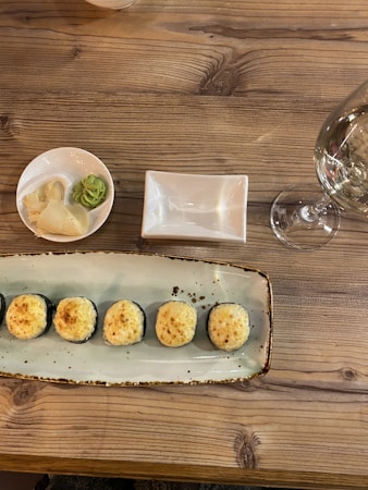A wooden table displays a plate with six pieces of sushi rolls, topped with a golden-brown baked crust. To the left is a small dish containing pickled ginger and wasabi. A glass of clear white wine sits on the right, next to an empty soy sauce dish.