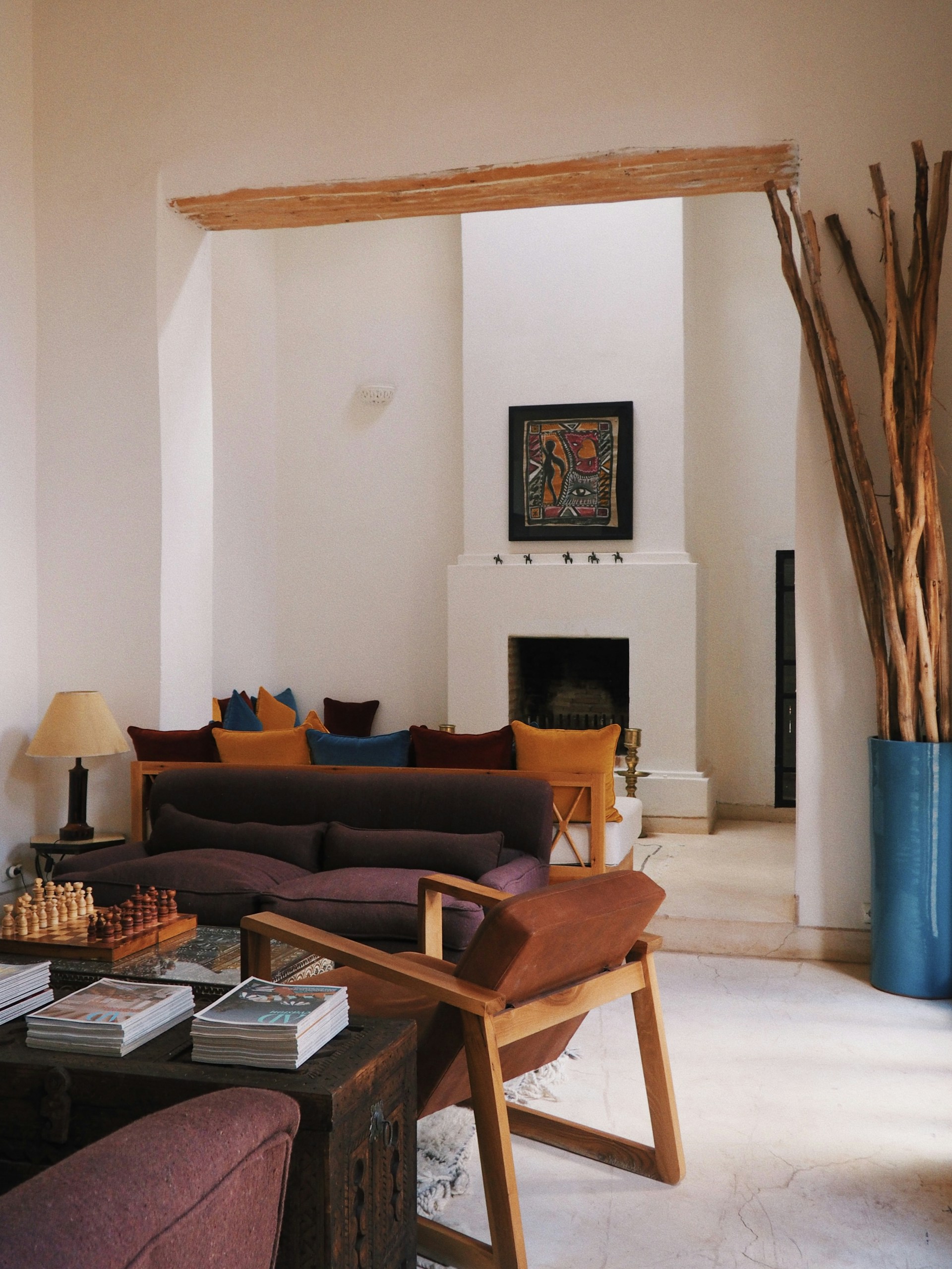 A cozy living room features a mix of modern and rustic elements. A plush maroon sofa and matching chairs are set around a dark wooden coffee table with a glass top, which holds a chess set and stacks of magazines. Tall dried branches are arranged in a large blue vase, adding an organic touch. In the background, colorful cushions adorn a wooden bench against a fireplace, with a vibrant abstract painting hanging above. The walls are painted in a soft neutral tone, complemented by natural light streaming in.