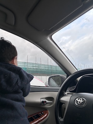 A child is seated inside a car, looking out the window. The interior shows the car's steering wheel, dashboard, and window controls. Outside, there appears to be a construction site with scaffolding covered in green netting.