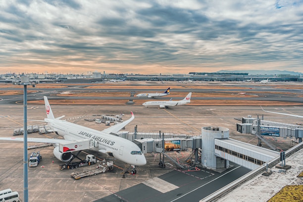 A bustling airport scene with several airplanes on the runway and at the gates. A Japan Airlines plane is parked at a gate, with boarding equipment and cargo vehicles around it. Another plane takes off in the background, with airport infrastructure visible under a cloudy sky.