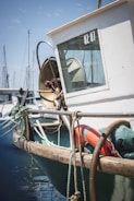 A fishing boat docked at a marina with ropes and a life buoy attached. The boat has a white cabin with a window displaying '02 01'. There are other boats and masts visible in the background.
