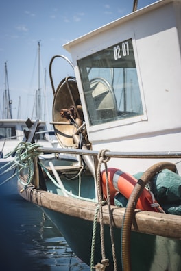 A fishing boat docked at a marina with ropes and a life buoy attached. The boat has a white cabin with a window displaying '02 01'. There are other boats and masts visible in the background.