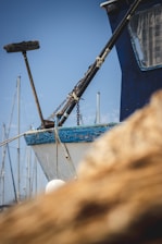 A close-up view of the front part of a blue and white boat docked at a marina. The boat has ropes and chains, and a mop-like object is attached to a bar. Sailboat masts are visible in the background against a clear blue sky. A blurred foreground suggests adjacent boats or equipment.