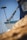 A close-up view of the front part of a blue and white boat docked at a marina. The boat has ropes and chains, and a mop-like object is attached to a bar. Sailboat masts are visible in the background against a clear blue sky. A blurred foreground suggests adjacent boats or equipment.