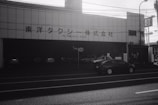 A black and white image of a building with Japanese text across the front. The signage suggests it could be a taxi company or garage. Several cars are parked in front of the building, and a dark-colored sedan is driving past on the street. Overhead wires and streetlights are visible, with shadows indicating daytime.