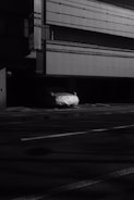A dimly lit urban scene featuring a parked car covered with a white protective sheet. The building in the background displays a grid-like pattern on its exterior surface.
