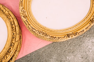 Ornate golden picture frames with intricate floral designs are placed partially on a pink surface and a gray textured background. The frames have an empty center, indicating they are not currently holding any artwork.