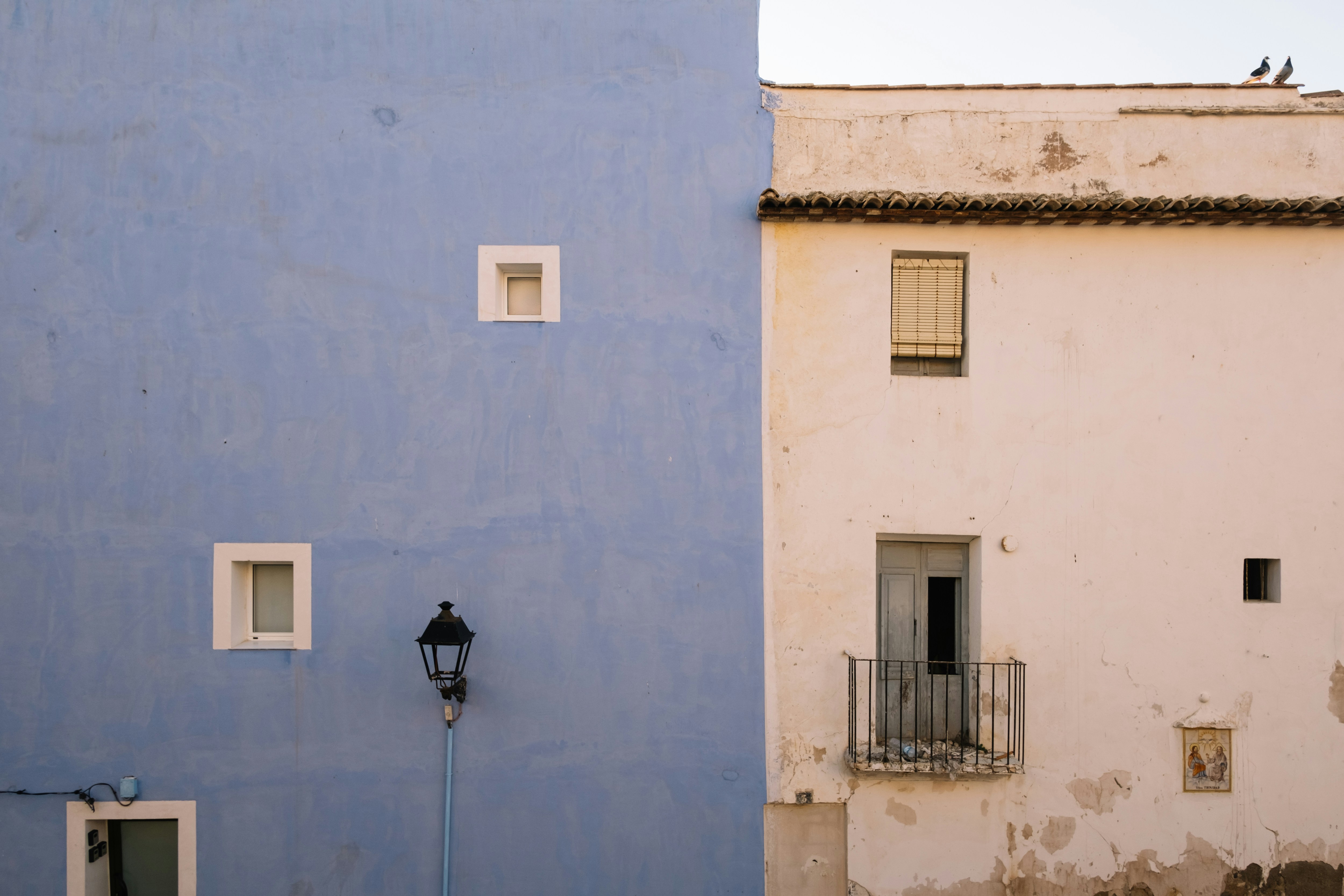 a blue building with two windows and a street light, A top view of the colorful houses in the Spanish village Villajoyosa. Beautiful white and blue building. Two pigeons sitting in the corner of a rooftop.