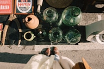 Various vintage items are laid out on a surface, including several large glass jars, a terracotta pot, a green ceramic pitcher, old wooden tools, and vintage beer advertisements. A person's feet in brown shoes and a white coat are visible at the bottom of the image, suggesting they are looking down at the items.