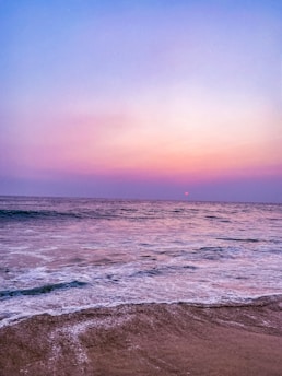 An ocean landscape at sunset with a calm sea and waves gently reaching the sandy shore. The sky is painted in hues of purple, pink, and blue, creating a tranquil and serene scene.