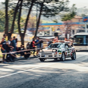 A rally car is speeding down a city street, surrounded by spectators who are blurred due to the motion of the vehicle. The car has racing decals and is captured in action, suggesting a high-speed event. Trees and urban elements are visible in the background, adding to the dynamic scene.