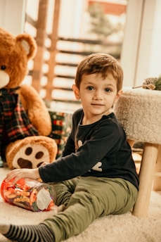 A young child is sitting on a soft carpet in a warmly lit room, holding a jar filled with colorful plastic letters. A large stuffed teddy bear wearing a plaid shirt is positioned behind the child. The setting appears cozy and playful, with soft furnishings and natural light coming from a window in the background.