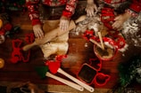 Two children with festive sweaters are engaged in baking activities. They are rolling out dough on a floured wooden surface. Cookie cutters in various shapes, including stars and gingerbread men, are scattered around. A red mixing bowl with wooden spoons and additional utensils is also present.