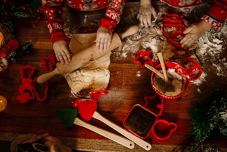 Two children with festive sweaters are engaged in baking activities. They are rolling out dough on a floured wooden surface. Cookie cutters in various shapes, including stars and gingerbread men, are scattered around. A red mixing bowl with wooden spoons and additional utensils is also present.
