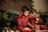 Two children wearing matching red Christmas sweaters with gingerbread men and snowflake patterns are engaged in a playful activity, possibly baking or playing with holiday-themed toys. The setting appears cozy, with a brick wall and soft pillows in the background. A small evergreen plant and red cups on the table contribute to a festive atmosphere.