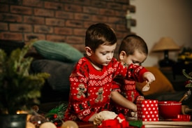 Two children wearing matching red Christmas sweaters with gingerbread men and snowflake patterns are engaged in a playful activity, possibly baking or playing with holiday-themed toys. The setting appears cozy, with a brick wall and soft pillows in the background. A small evergreen plant and red cups on the table contribute to a festive atmosphere.