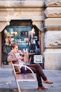 A man in a brown suit is sitting on a bench on a sidewalk, looking at his phone. He is in front of a clothing store with mannequins displaying suits in the window. The architecture features ornate stonework.