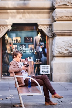 A man in a brown suit is sitting on a bench on a sidewalk, looking at his phone. He is in front of a clothing store with mannequins displaying suits in the window. The architecture features ornate stonework.