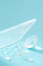 A delicate gold necklace with a pendant is elegantly suspended above a textured, translucent panel. Nearby, a large, faceted crystal and a few scattered white petals rest against a soft aqua background, creating a serene and balanced composition.