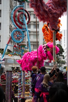 A vibrant street scene featuring a colorful and festive display with performers engaged in a traditional lion dance. The performers are handling a large pink lion costume while an elaborate, colorful structure is visible in the background. A crowd watches the performance, which is adorned with decorations and confetti.