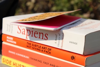A stack of books is shown with titles visible, including 'Sapiens' by Yuval Noah Harari, 'The Subtle Art of Not Giving a F*ck' by Mark Manson, 'Thinking, Fast and Slow' by Daniel Kahneman, and 'Side Hustle'. On top of the stack is a pink notebook with a yellow and black cover partially in view. The books are positioned outdoors with sunlight casting shadows.