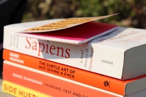 A stack of books is shown with titles visible, including 'Sapiens' by Yuval Noah Harari, 'The Subtle Art of Not Giving a F*ck' by Mark Manson, 'Thinking, Fast and Slow' by Daniel Kahneman, and 'Side Hustle'. On top of the stack is a pink notebook with a yellow and black cover partially in view. The books are positioned outdoors with sunlight casting shadows.