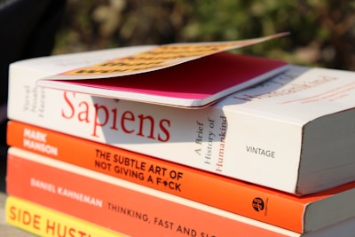 A stack of books is shown with titles visible, including 'Sapiens' by Yuval Noah Harari, 'The Subtle Art of Not Giving a F*ck' by Mark Manson, 'Thinking, Fast and Slow' by Daniel Kahneman, and 'Side Hustle'. On top of the stack is a pink notebook with a yellow and black cover partially in view. The books are positioned outdoors with sunlight casting shadows.