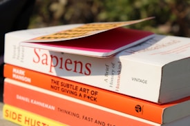 A stack of books is shown with titles visible, including 'Sapiens' by Yuval Noah Harari, 'The Subtle Art of Not Giving a F*ck' by Mark Manson, 'Thinking, Fast and Slow' by Daniel Kahneman, and 'Side Hustle'. On top of the stack is a pink notebook with a yellow and black cover partially in view. The books are positioned outdoors with sunlight casting shadows.