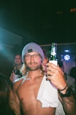 A man with long hair and a beard is wearing glasses and a gray beanie, holding a bottle of beer in a lively party setting. He is shirtless, has tattoos on his arm, and appears confident and relaxed. Neon lights in the background suggest a nightclub or bar atmosphere.