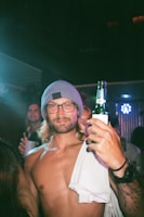 A man with long hair and a beard is wearing glasses and a gray beanie, holding a bottle of beer in a lively party setting. He is shirtless, has tattoos on his arm, and appears confident and relaxed. Neon lights in the background suggest a nightclub or bar atmosphere.