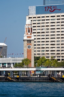 A historic clock tower stands prominently in front of a modern high-rise building that displays a large sign with the numbers '175' and Chinese characters. The foreground features a waterfront area with palm trees, flags, and a promenade where people are walking. The sky is clear blue and the water is a deep shade of blue.
