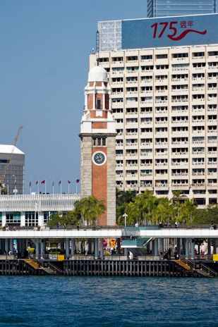 A historic clock tower stands prominently in front of a modern high-rise building that displays a large sign with the numbers '175' and Chinese characters. The foreground features a waterfront area with palm trees, flags, and a promenade where people are walking. The sky is clear blue and the water is a deep shade of blue.