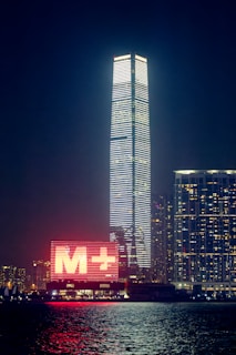 A night-time cityscape featuring a tall skyscraper illuminated with horizontal white lights. To the left of the skyscraper, there is a large digital sign with a red 'M+' displayed prominently. The scene also includes other buildings with numerous lit windows, and the foreground includes a body of water reflecting the city lights.