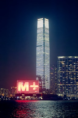 A night-time cityscape featuring a tall skyscraper illuminated with horizontal white lights. To the left of the skyscraper, there is a large digital sign with a red 'M+' displayed prominently. The scene also includes other buildings with numerous lit windows, and the foreground includes a body of water reflecting the city lights.