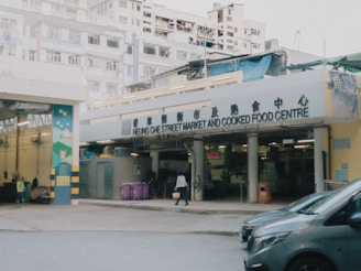 A street market sign reads 'Heung Che Street Market and Cooked Food Centre' with a city apartment building in the background. Pedestrians are seen entering and exiting the market area. Trash bins and vehicles are parked nearby, and the overall scene looks overcast and slightly hazy.