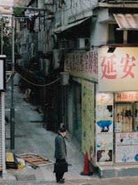 An urban alleyway scene featuring a man walking with a briefcase past a storefront with large Chinese signage. The alley is bordered by older buildings, some with visible air conditioning units and hanging laundry. A traffic cone and some construction materials lie near the entrance to the alley.