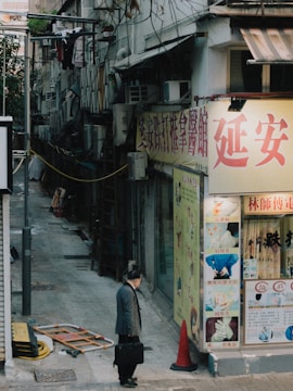 An urban alleyway scene featuring a man walking with a briefcase past a storefront with large Chinese signage. The alley is bordered by older buildings, some with visible air conditioning units and hanging laundry. A traffic cone and some construction materials lie near the entrance to the alley.