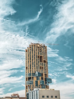 A modern high-rise building with a unique, cylindrical architecture stands against a backdrop of a clear blue sky. The structure features a crane on top, which suggests ongoing construction or maintenance work. The façade is predominantly composed of large, glossy windows and beige stone or concrete elements.