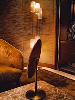A warmly lit interior features a modern floor lamp with multiple cylindrical shades, casting a soft light against a textured wall. The room includes a plush armchair with rich fabric and a sleek wooden table with a marble top. The overall design aesthetics are sophisticated and cozy.